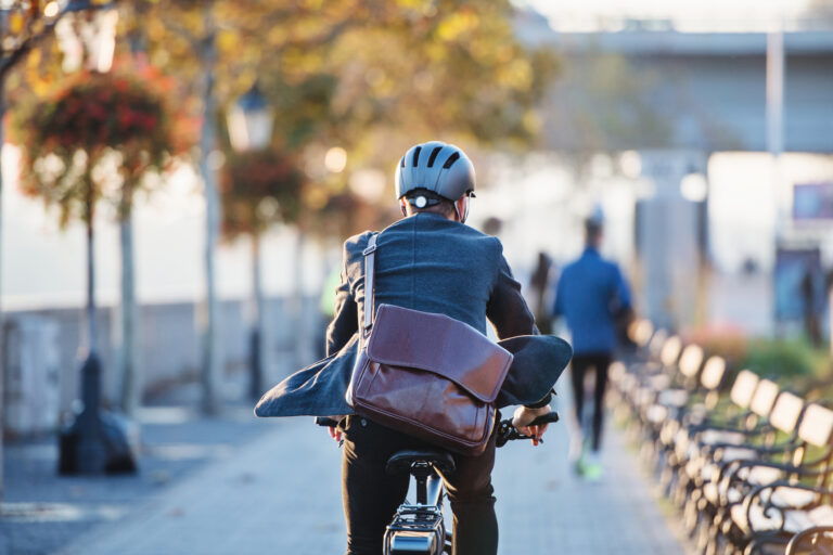 Person riding a bicycle away from the camera on a city street with autumn foliage in the background.