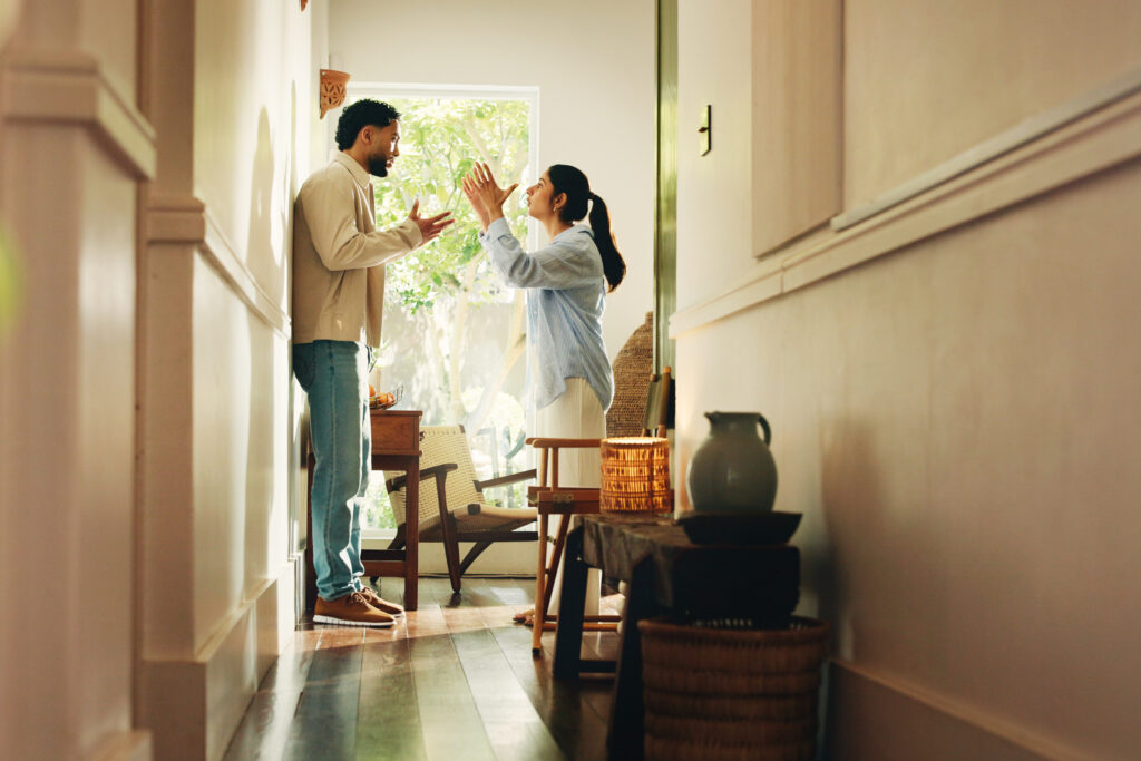 Two people in a narrow hallway facing each other, gesturing with raised hands as they talk near a window.