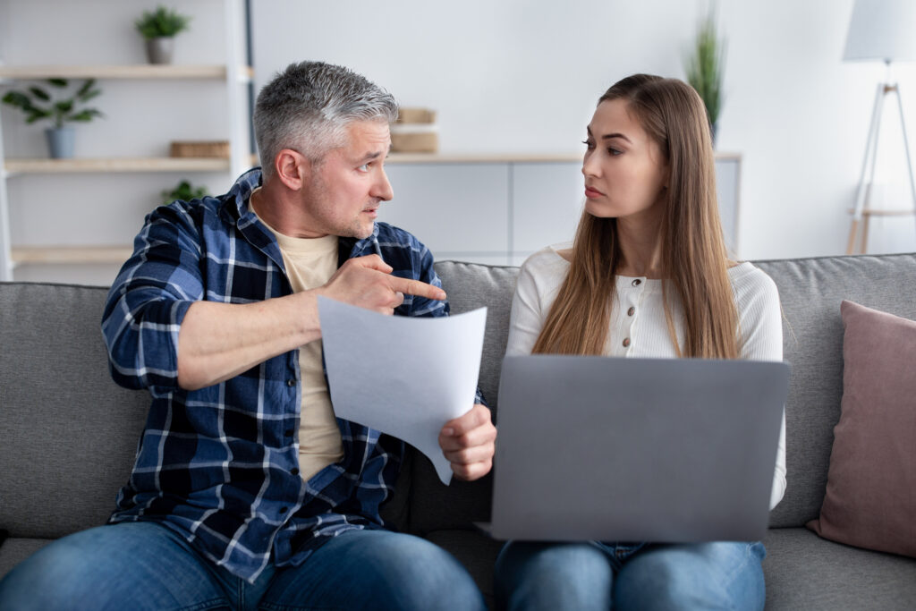 Man and woman seated on a sofa, reviewing papers with a laptop nearby in a living room setting.
