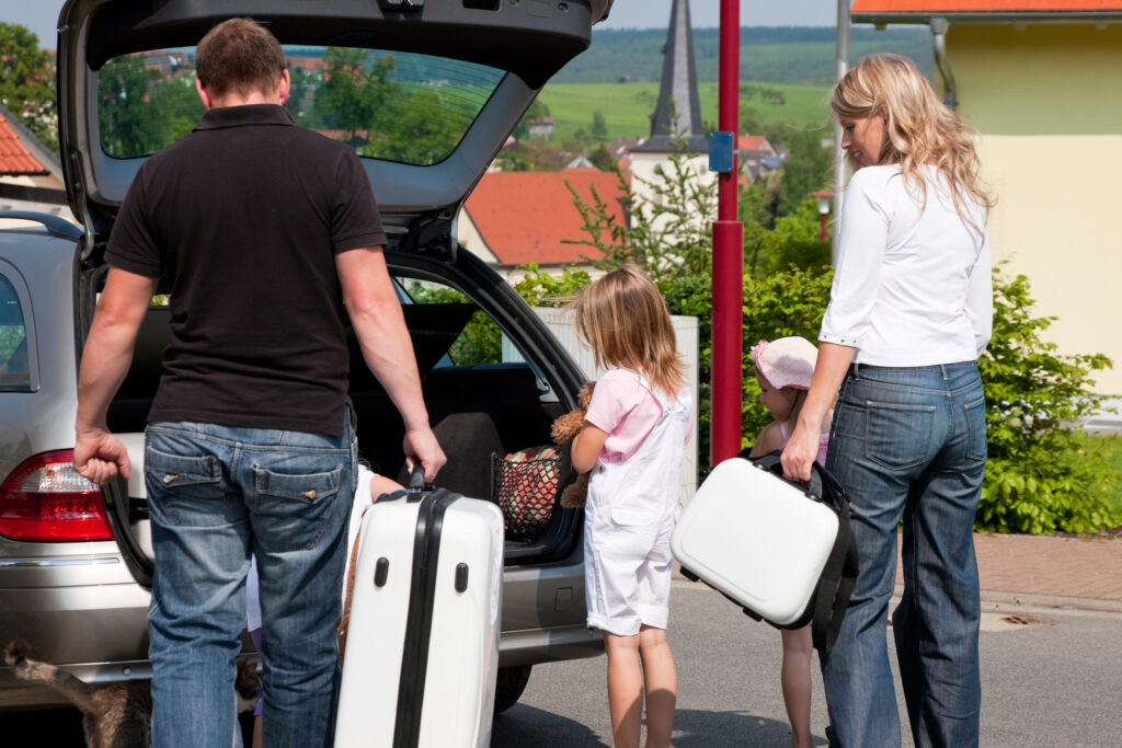 Family loading white suitcases from the open trunk of a silver SUV, with a small dog nearby in a parking area.